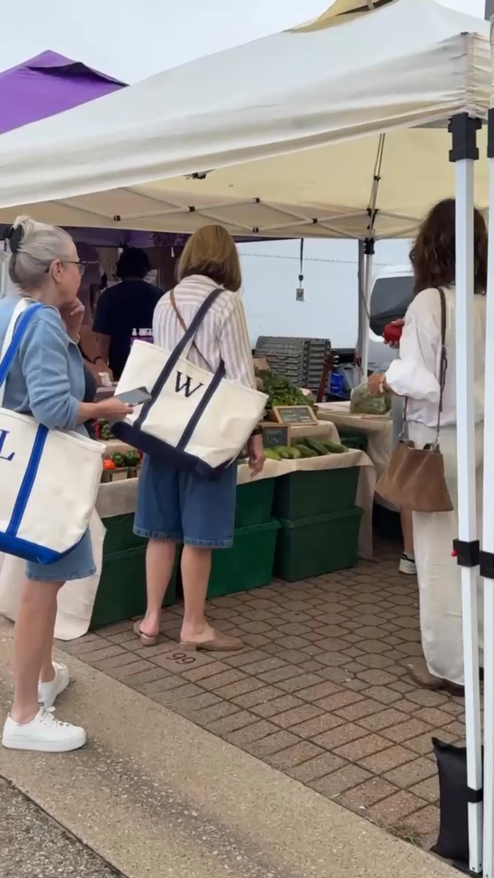 Three women with tote bags shop for produce at the Found Cottage Mercantile Market under a white tent.