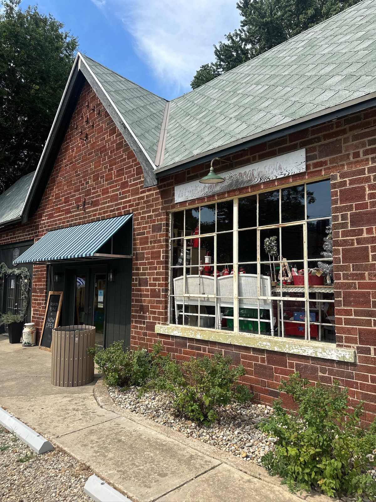 Red brick building with large windows, green awning, and found cottage mercantile market items displayed in the window.