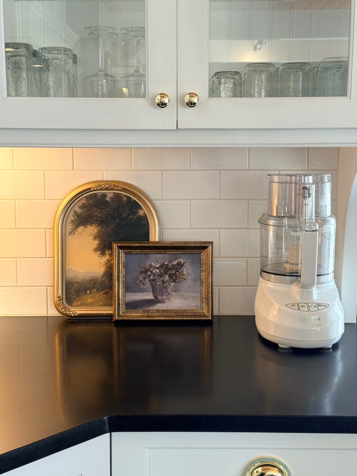 Two framed paintings and a food processor add charm to this white kitchen with muted fall decor on the black countertop.