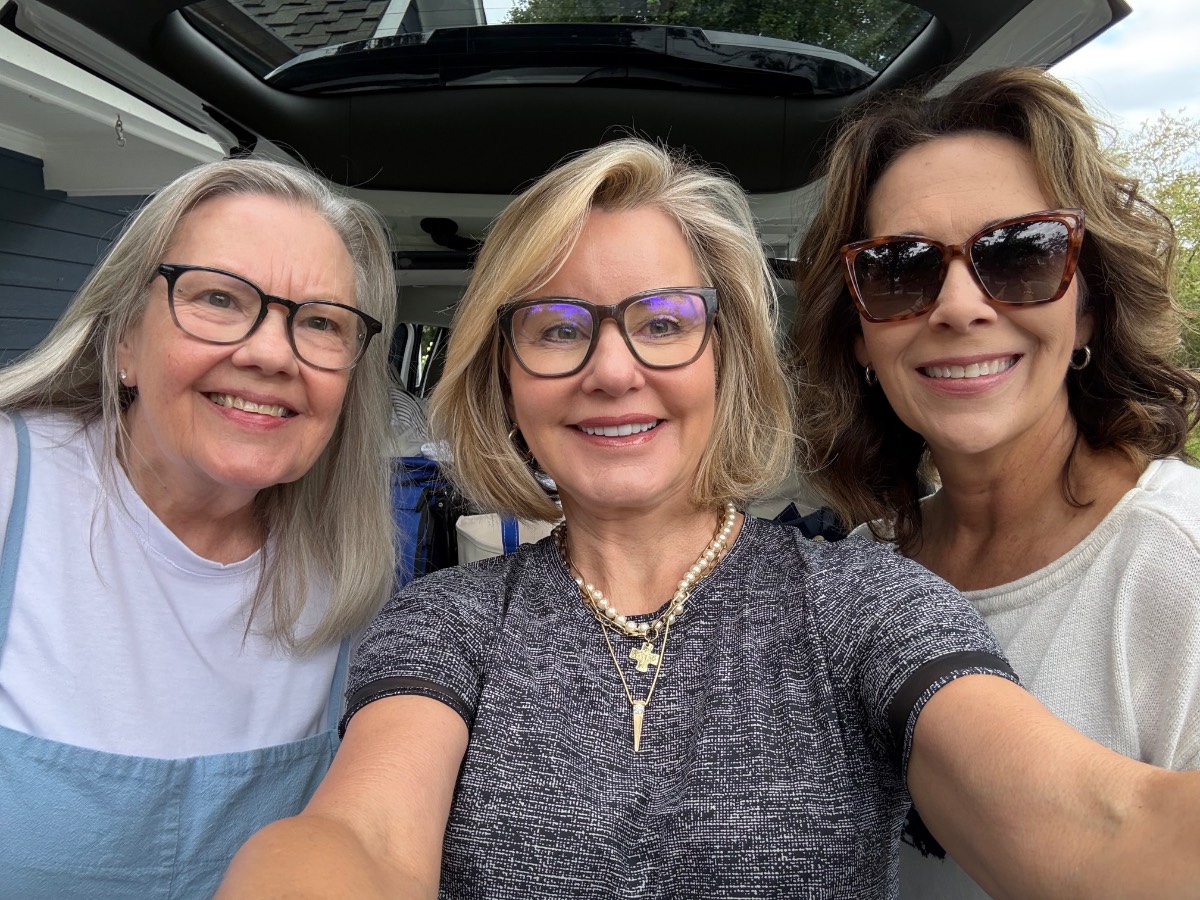 Three smiling women, all in the latest casual clothes and glasses, pose for a selfie outdoors near an open car trunk.