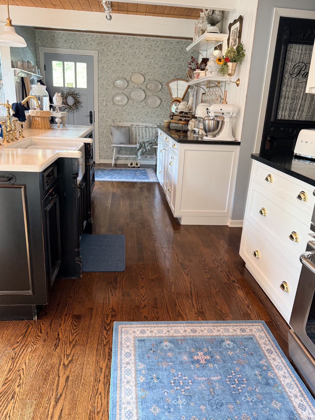 Bright kitchen with wooden floors, blue rugs, white cabinets, and cranberry decor displayed on a patterned wall.