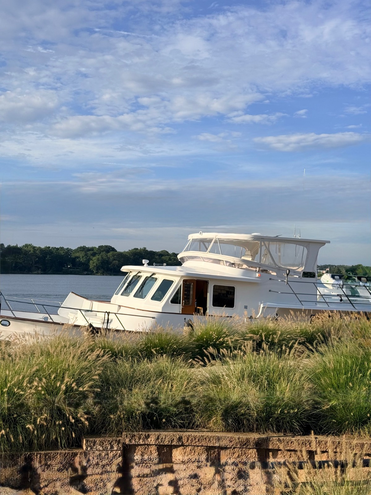 A white boat, featuring the latest design, is docked by a lake with green grass and blue sky in the background.