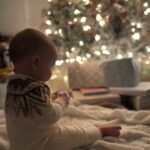 A baby in a latest patterned sweater sits on a blanket in front of a glowing Christmas tree with wrapped gifts.