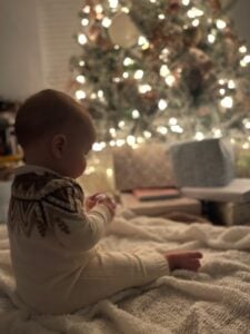 A baby in a latest patterned sweater sits on a blanket in front of a glowing Christmas tree with wrapped gifts.