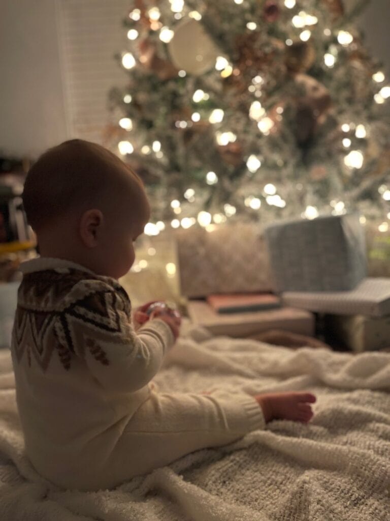 A baby in a latest patterned sweater sits on a blanket in front of a glowing Christmas tree with wrapped gifts.