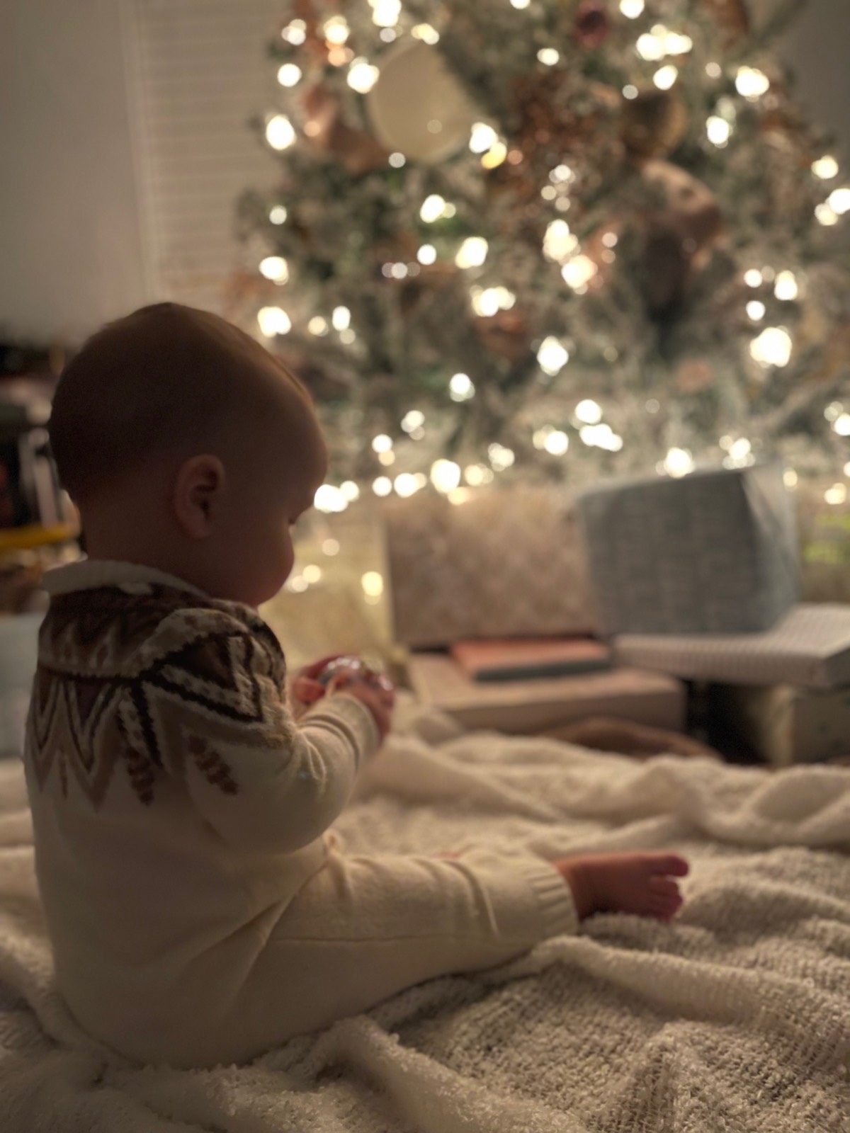 A baby in a latest patterned sweater sits on a blanket in front of a glowing Christmas tree with wrapped gifts.