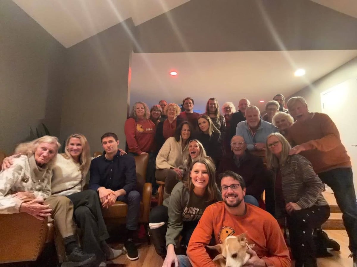 A group poses for a Thanksgiving hosting photo in a living room, with a man holding a small dog in the foreground.