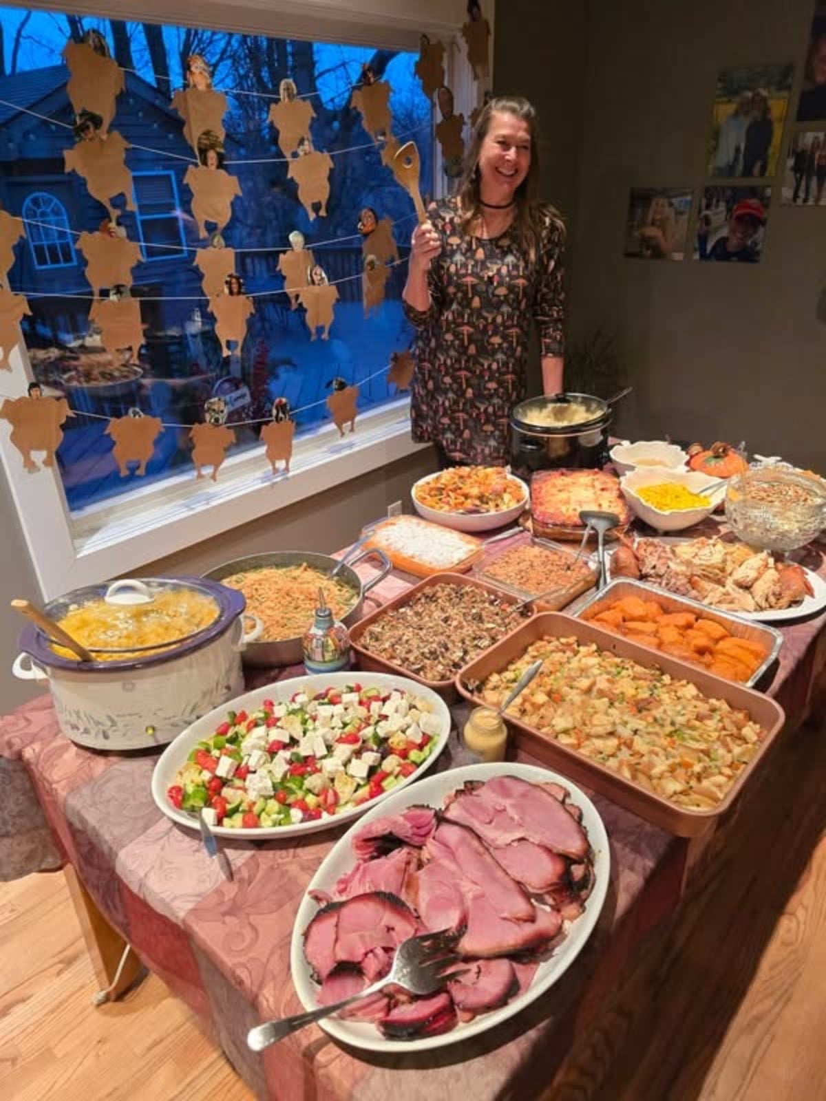 A woman holding a wooden spoon stands behind a table filled with Thanksgiving dishes, perfect for hosting Thanksgiving.