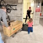 A man pushes a dolly of boxes toward a woman and child standing by a Christmas tree lit with firecracker lights.