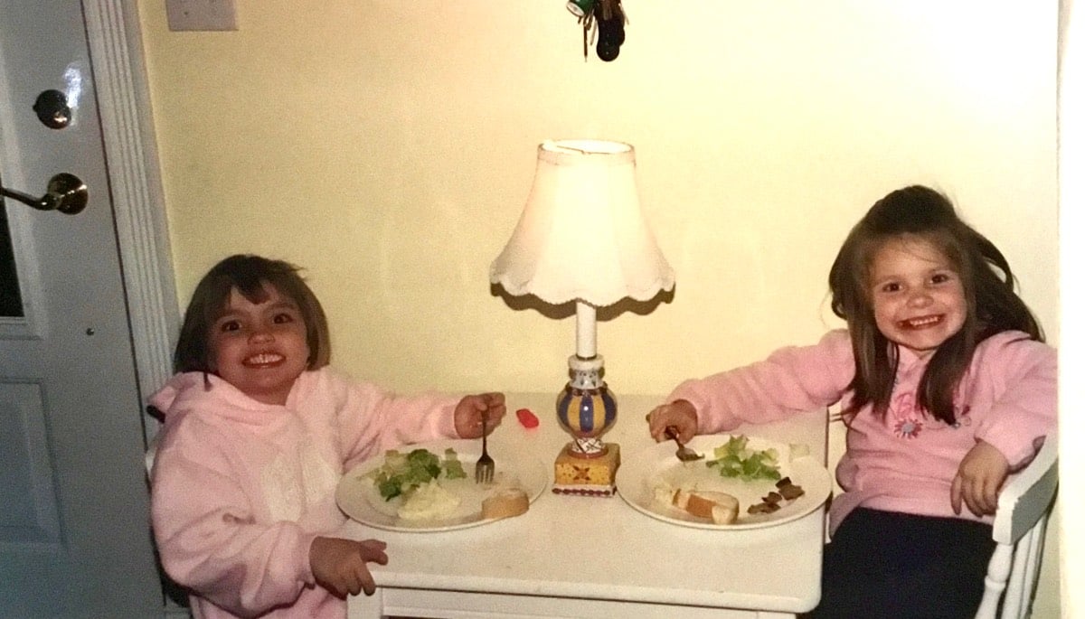 Two young girls in pink sweatshirts enjoy a small house Thanksgiving dinner at a table with a lamp between them.