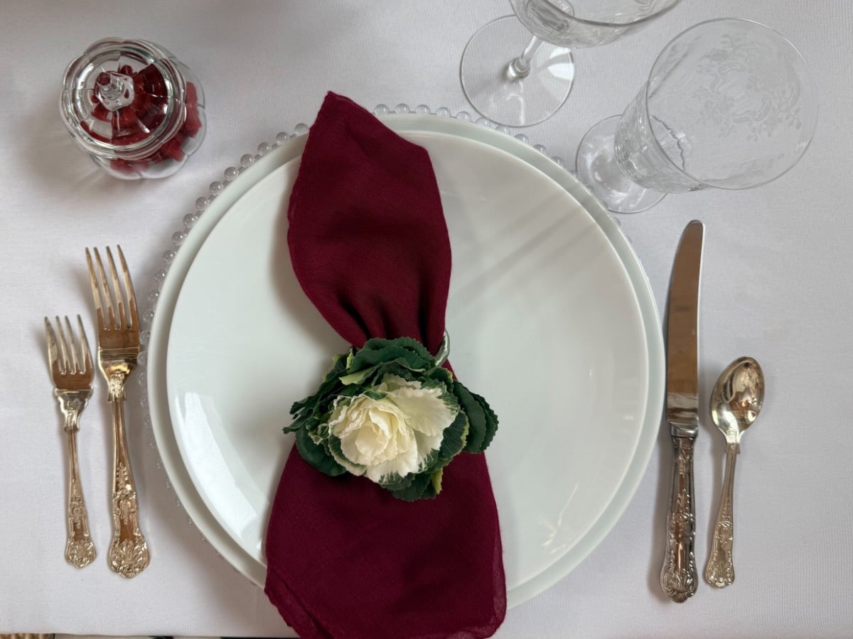A Thanksgiving place setting with white plates, a dark red napkin in a floral ring, silverware, and two glasses.