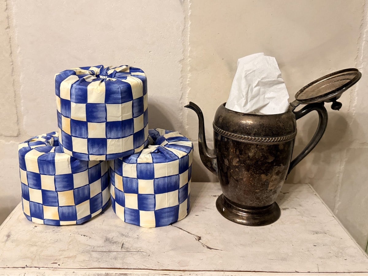 Three blue and white checkered toilet paper rolls beside an antique silver teapot holding a tissue for Thanksgiving hosting.
