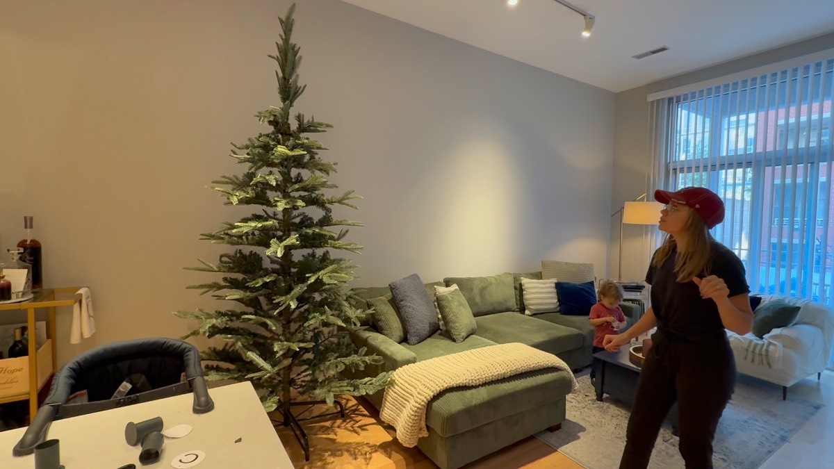 A woman stands in a living room near an undecorated artificial Christmas tree with firecracker lights and a green sectional.