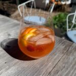 A glass of orange cocktail with ice, lemon, and a berry on a wooden table set for an outdoor fall dinner party.