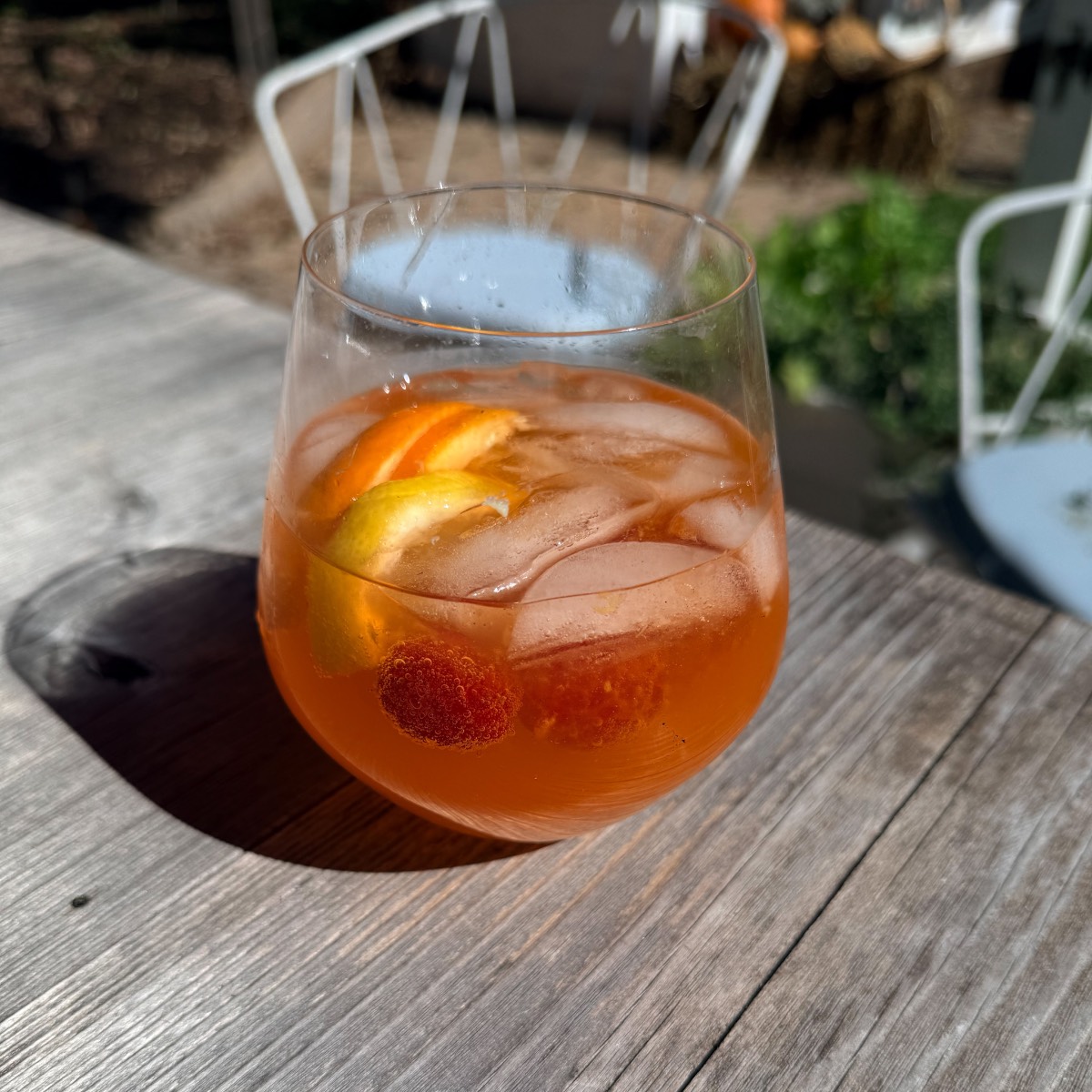 A glass of orange cocktail with ice, lemon, and a berry on a wooden table set for an outdoor fall dinner party.