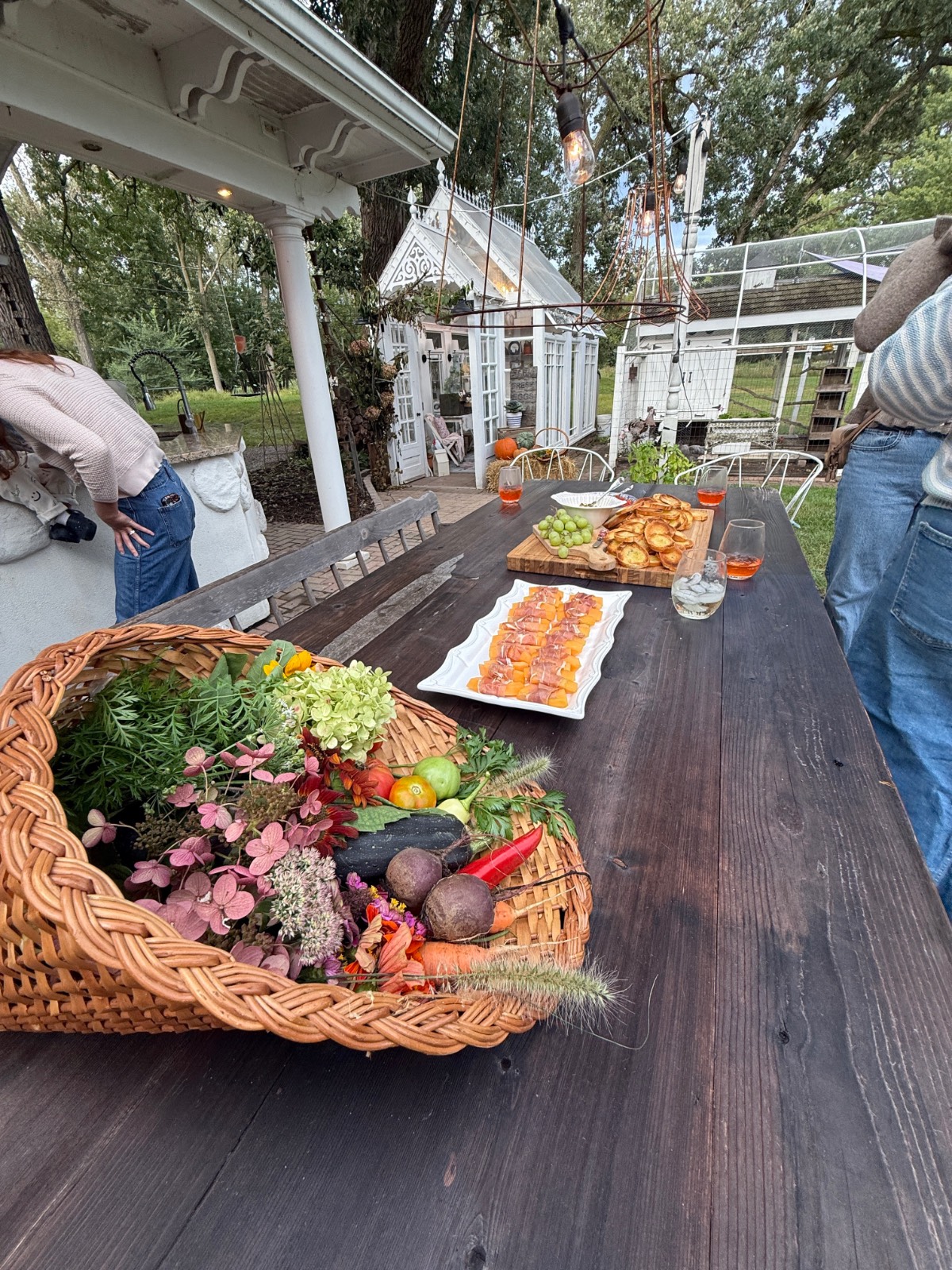 A rustic outdoor table set for a fall dinner party, with fresh produce, appetizers, drinks, and guests mingling nearby.