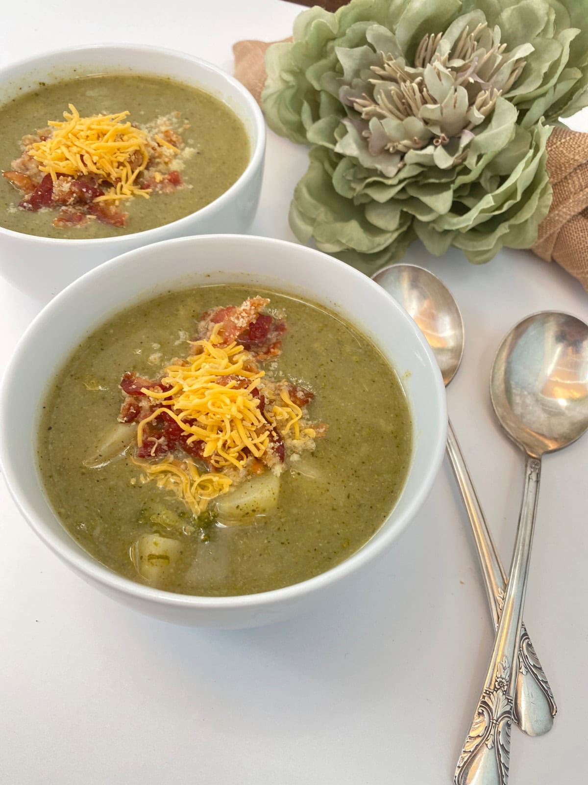 Two bowls of green vegetable soup topped with cheddar and bacon for your shopping list, served with two spoons.