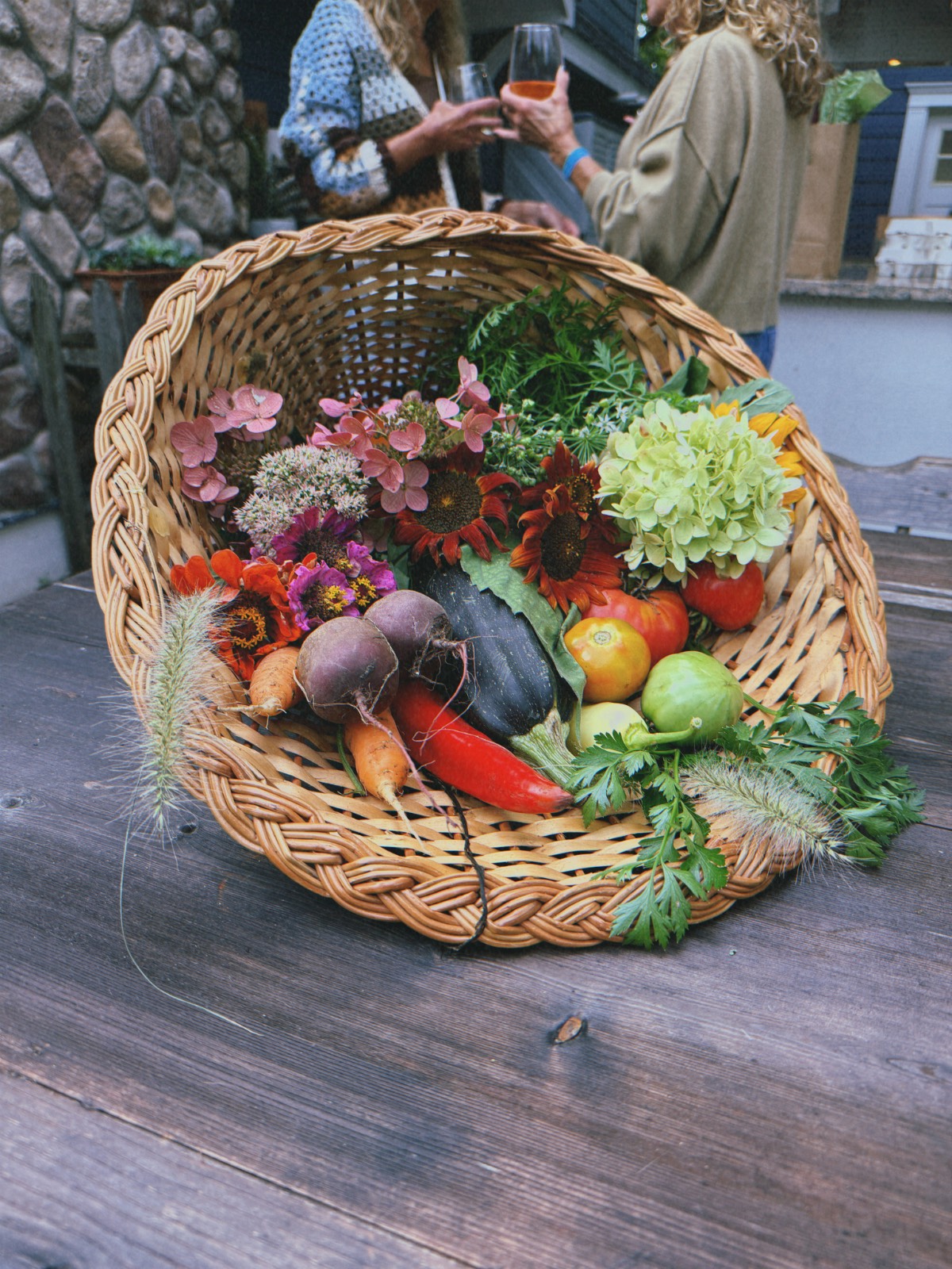 A woven basket spills flowers, veggies, and herbs as the host chats at an outdoor fall dinner party in the background.