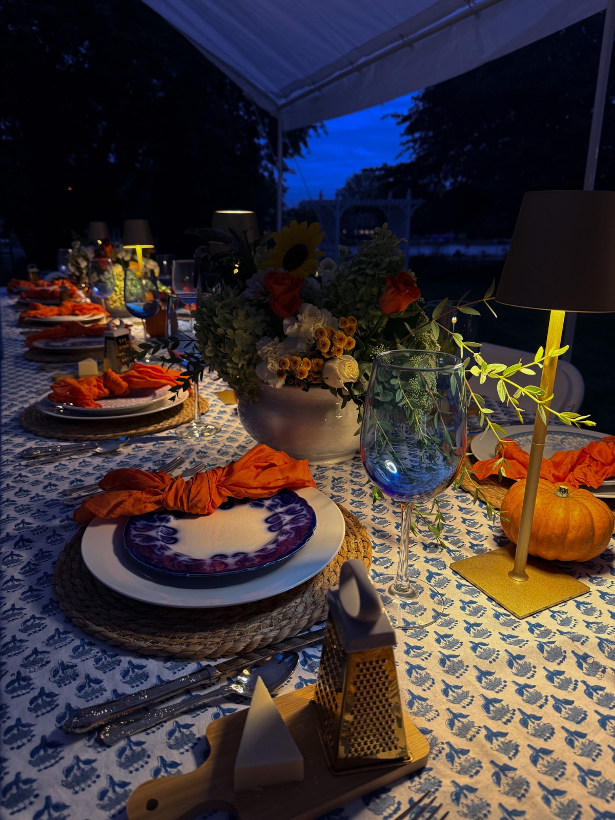 Elegant outdoor fall dinner party table at dusk with flowers, pumpkins, lamps, and cheese grater on a patterned cloth.