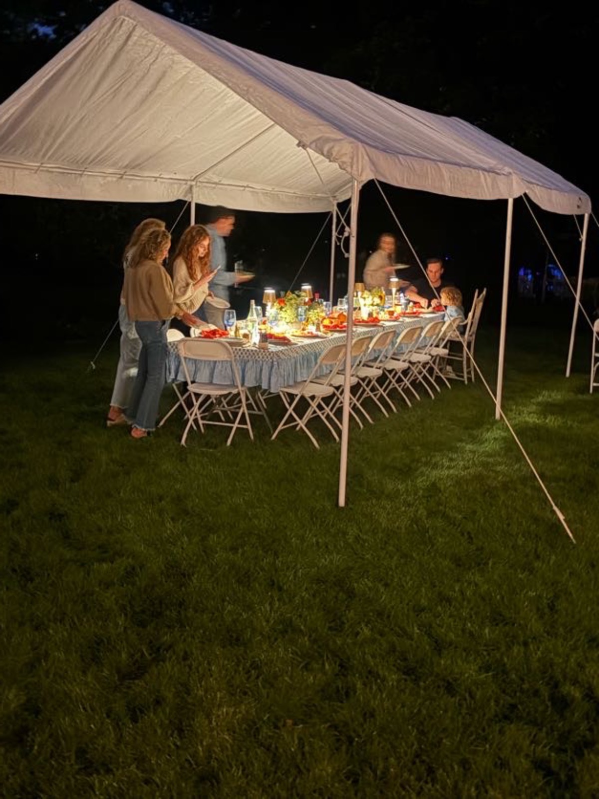 Guests gather at a long table under a white canopy for an elegant outdoor fall dinner party on the grassy lawn.