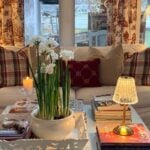 A living room coffee table featuring a pot of white narcissus flowers, a small lamp, and several stacked books.