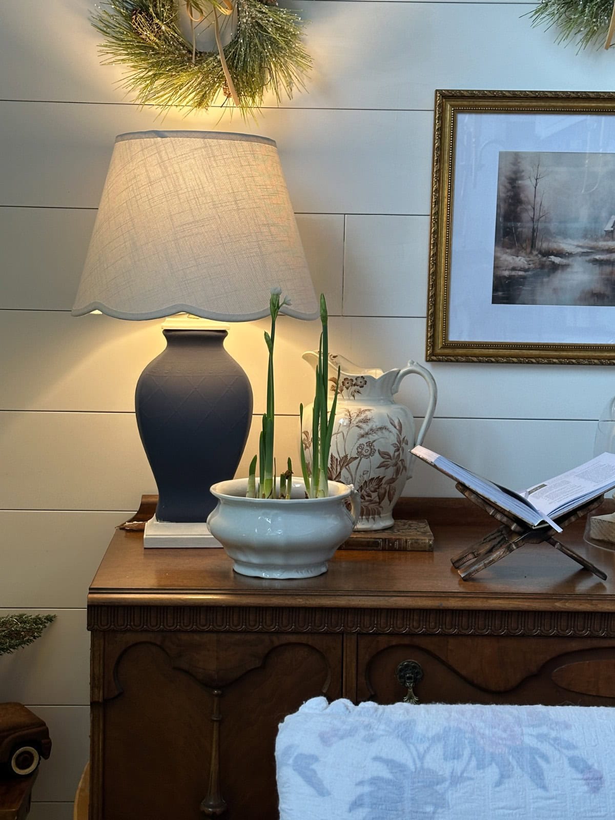 A lamp, a ceramic planter with sprouting bulbs, and an open book on a wooden stand sit on a wooden cabinet.