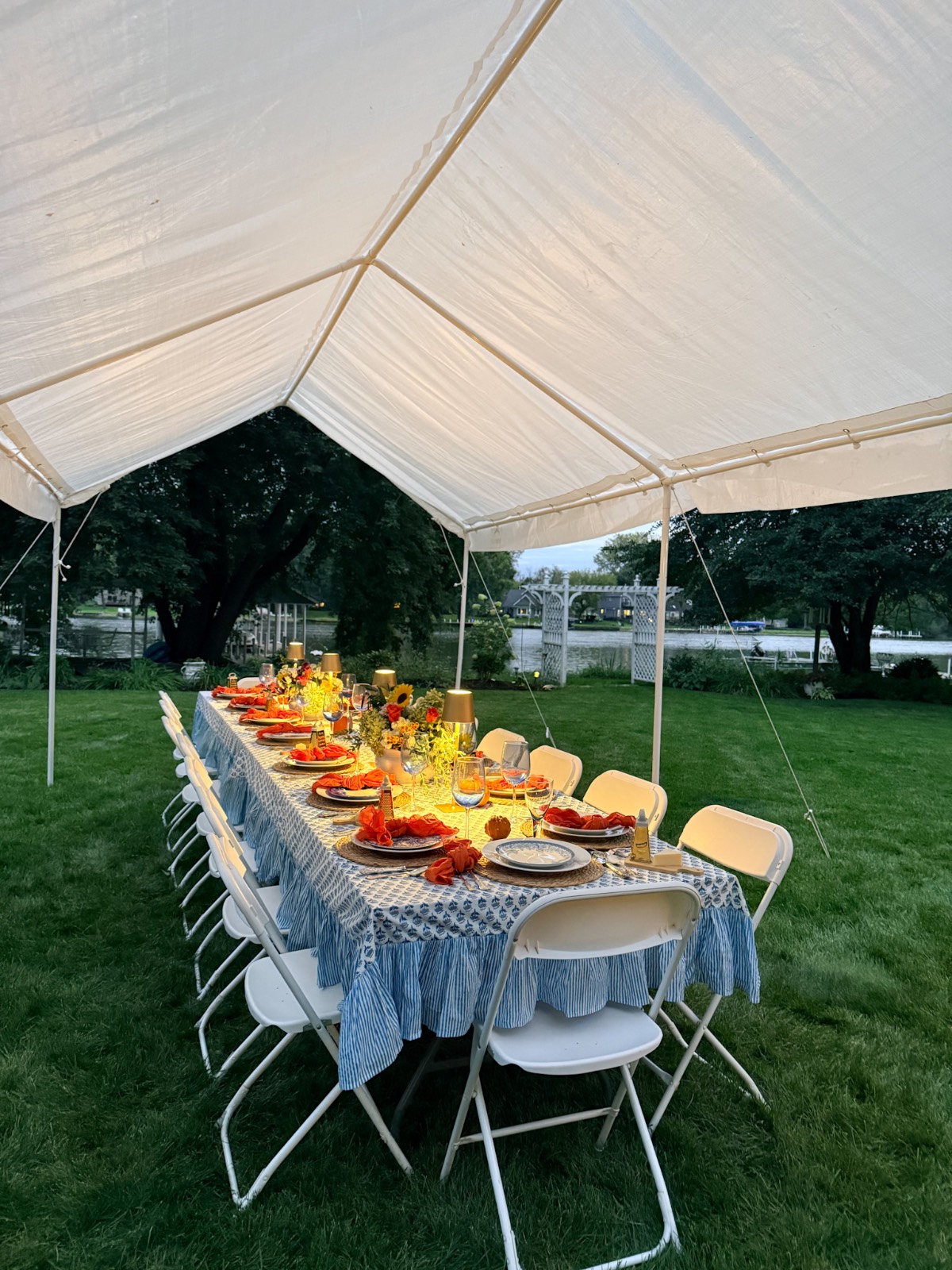 A long table with striped cloth set for a fall dinner party under a white canopy on a green lawn with folding chairs.