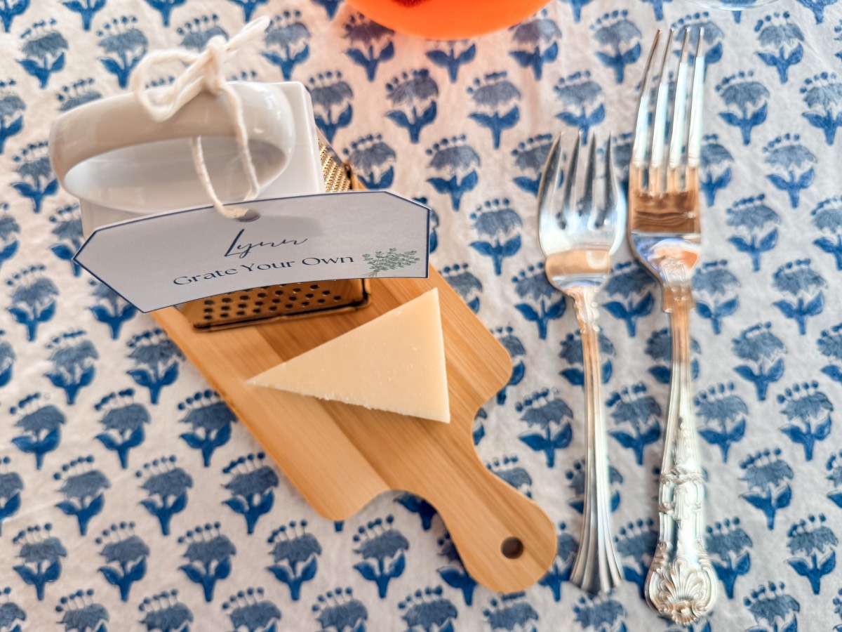 A small cheese grater, a wedge of cheese, and silver forks on a floral tablecloth for an outdoor fall dinner party.