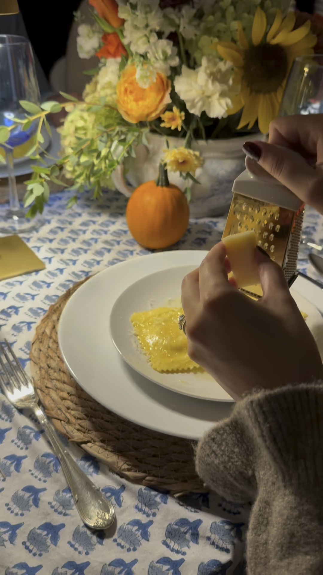 Hands grating cheese over ravioli on a white plate at an outdoor fall dinner party, with a pumpkin and flowers nearby.