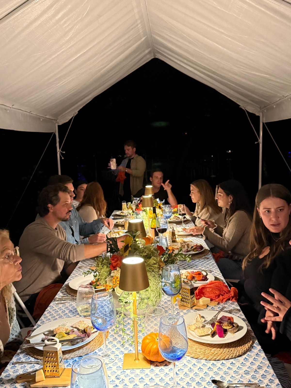 A group enjoys a festive fall dinner party outdoors at night, gathered around a long table beneath a white tent.