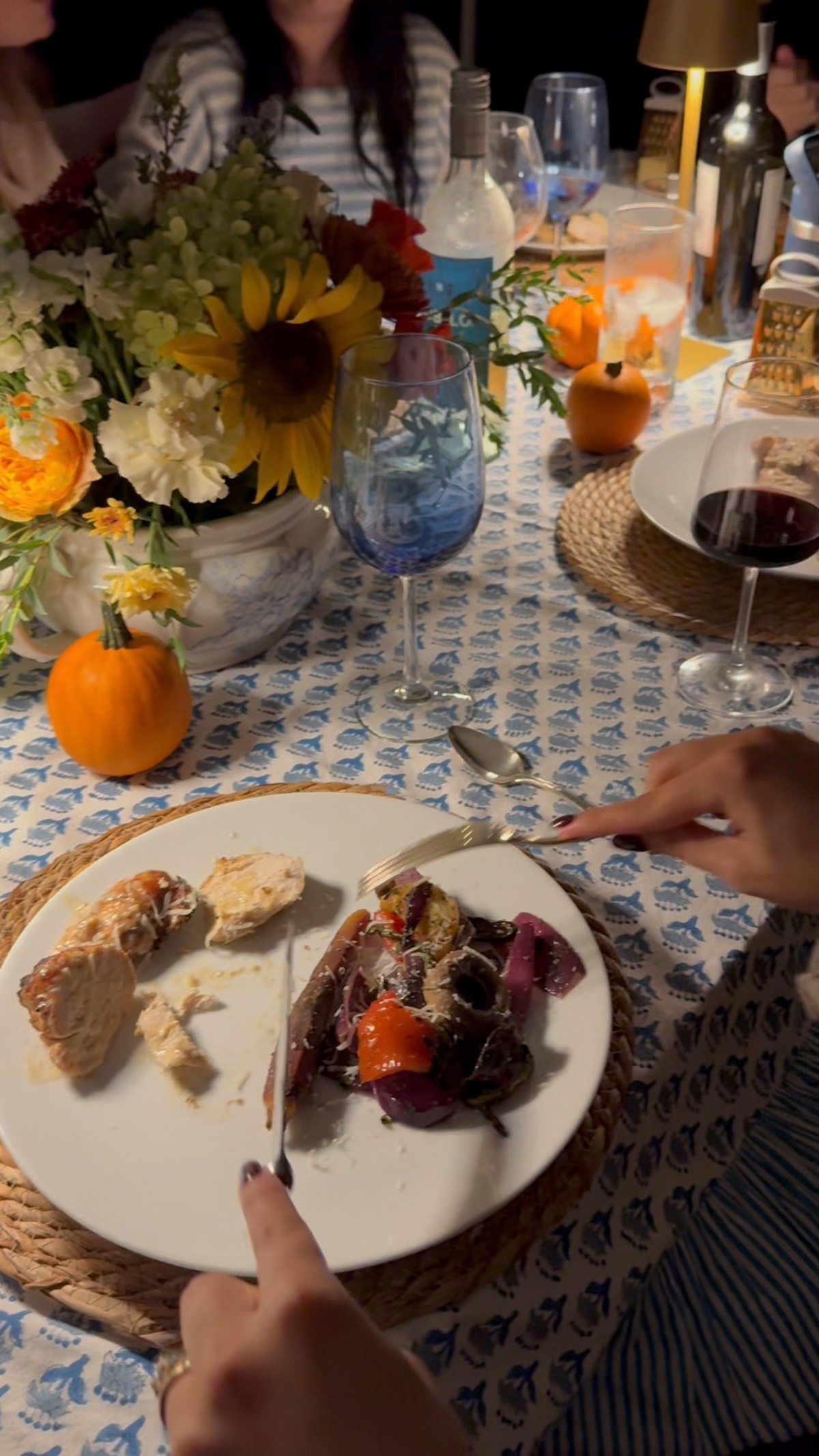 A person cuts food at an outdoor fall dinner party table set with flowers, mini pumpkins, bread, and wine.