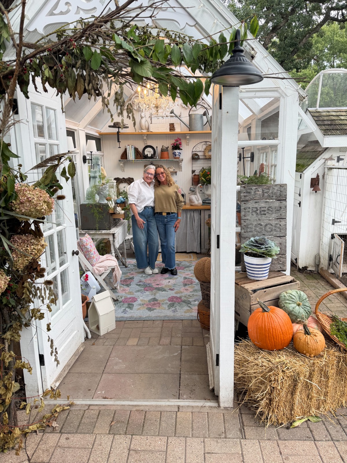 Two people smile inside a cozy garden shed, ready to host a charming outdoor fall dinner party amid pumpkins and plants.
