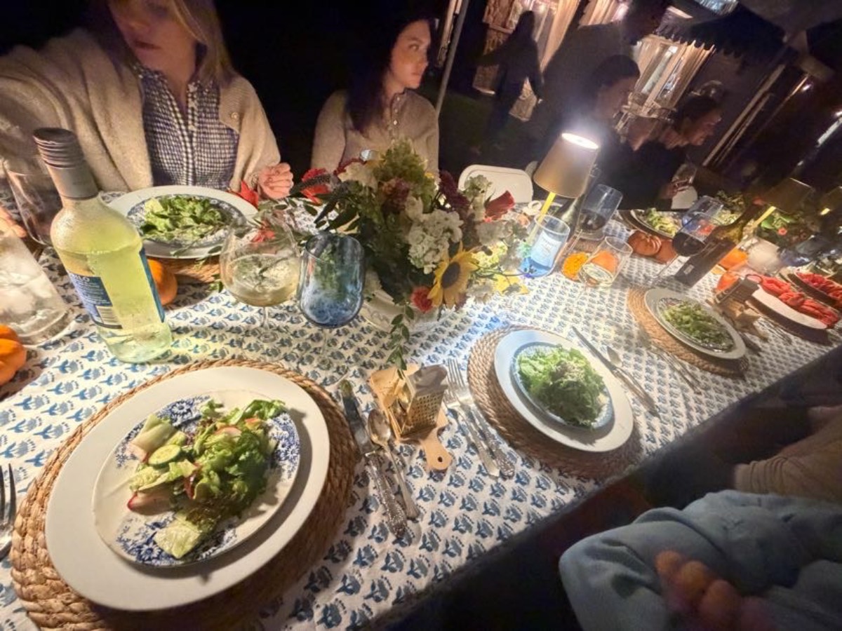 People enjoy a cozy fall dinner party at night, sharing salad and drinks on a patterned tablecloth outdoors.