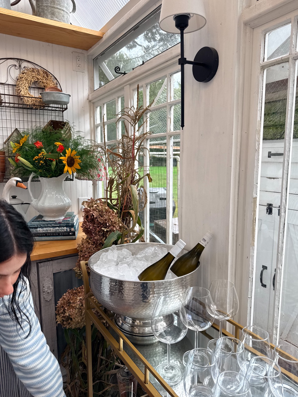 Woman beside a cart with wine bottles, glasses, and flowers—perfect for an outdoor fall dinner party by large windows.